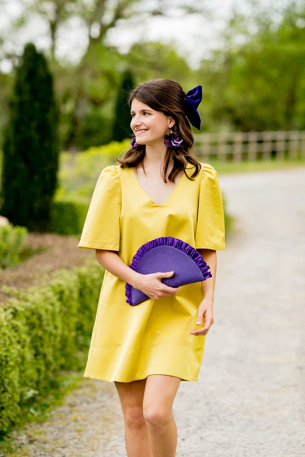 Jeune femme habillée de jaune portant une pochette violette de forme demi-lune dont dépassent des volants violets. Créé par Bellemond.