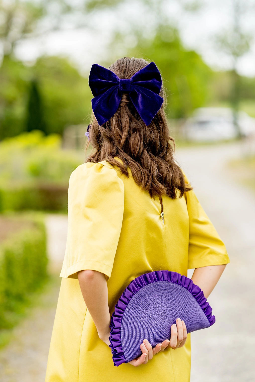 Jeune femme habillée de jaune et se tenant les mains dans le dos portant une pochette en demi-lune de couleur violette dont dépassent des fronces. Créé par Bellemond.