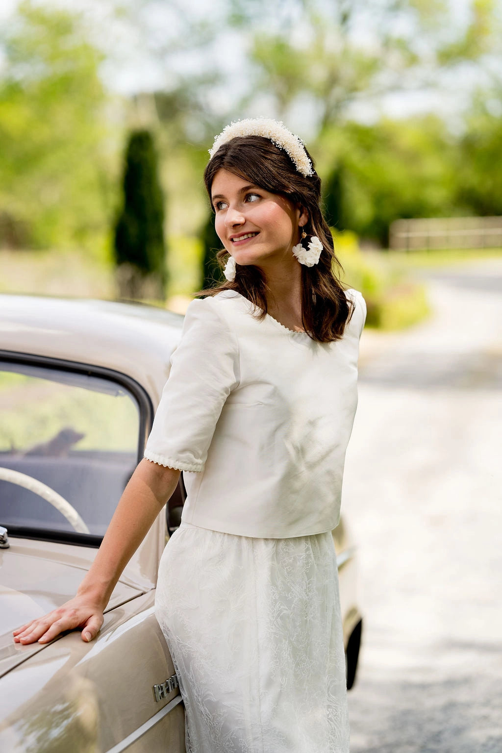 Mariée s'appuyant sur une voiture ancienne portant dans ses cheveux un serre-tête composé de mini gypsophile ivoire. Créé par Bellemond.
