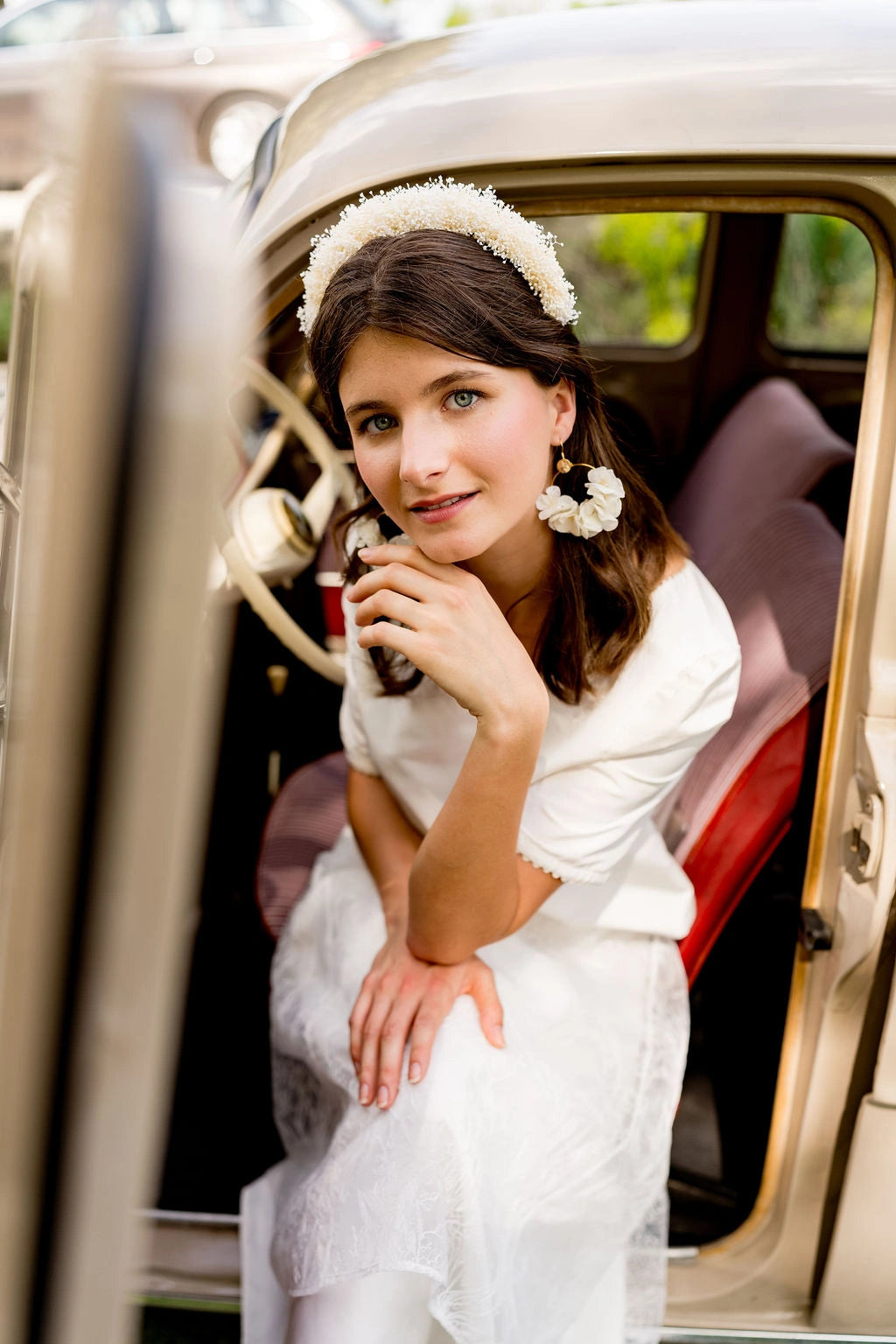 Mariée assise dans une voiture ancienne portant un serre-tête composé de mini gypsophile blanc dans les cheveux. Créé par Bellemond. 