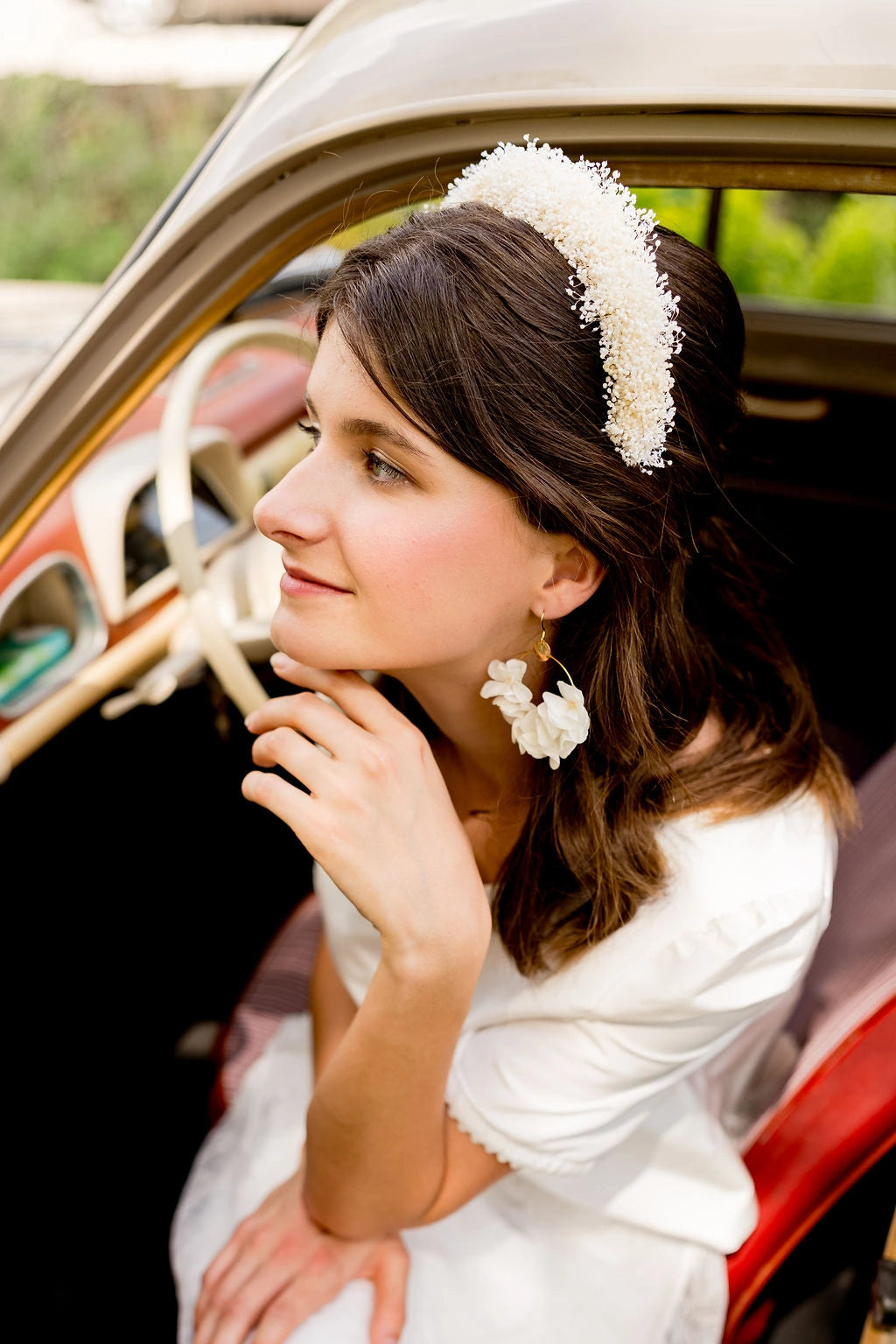 Mariée assise sur le siège avant d'une voiture ancienne portant dans les cheveux un serre-tête composé de mini gypsophile ivoire. Créé par Bellemond. 