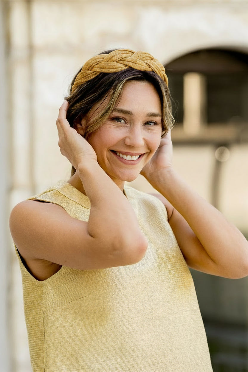 Jeune femme portant les mains à la tête ou se trouve dans les cheveux un serre-tête en sinamay doré tressé. Créé par Bellemond.