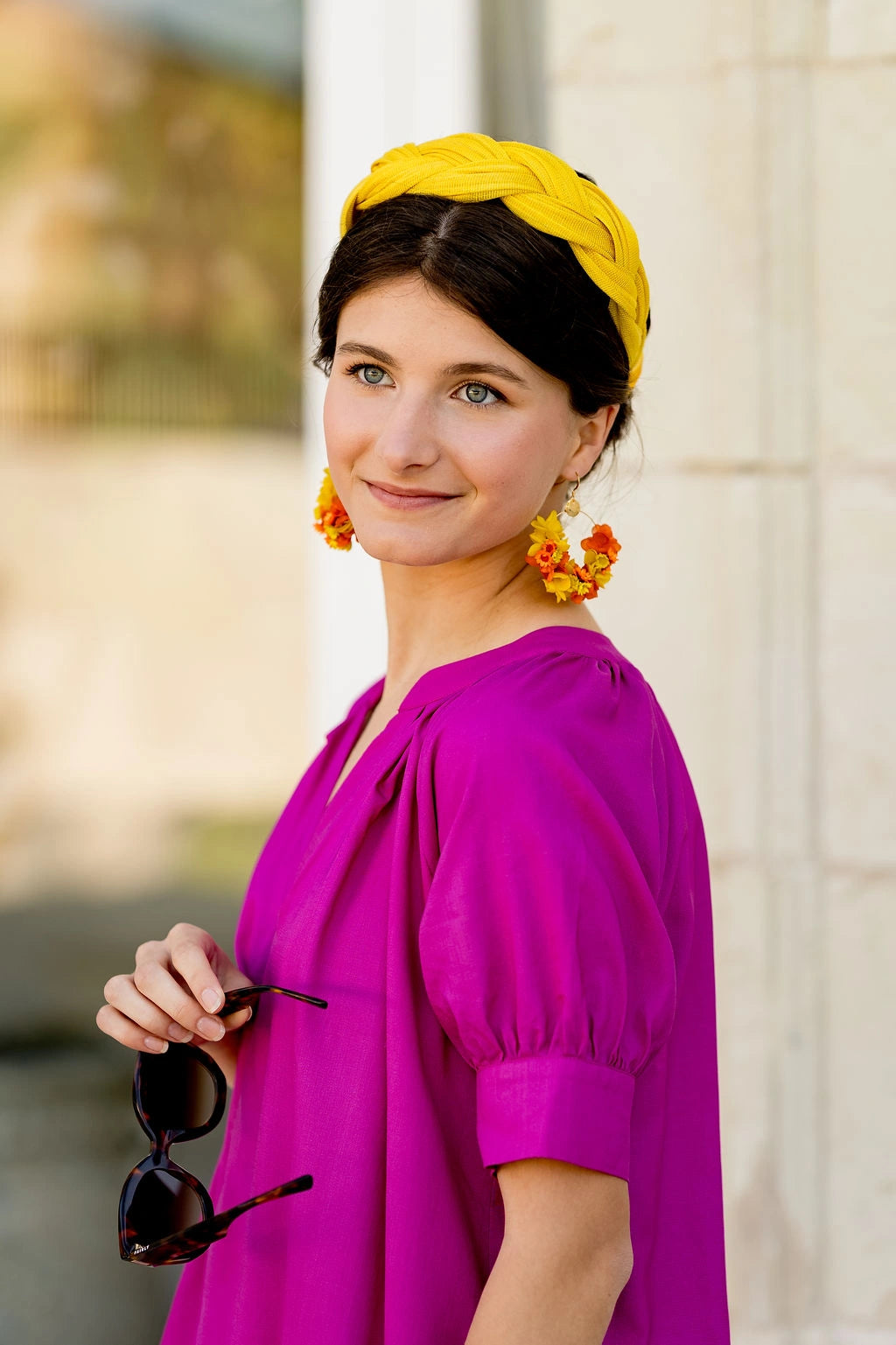 Jeune femme habillée de fuchsia et tenant des lunettes de soleil. Elle porte dans les cheveux un serre-tête de couleur jaune tressé en sinamay. Créé par Bellemond. 